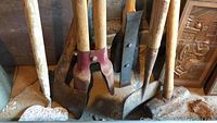 Close view of seven garden and hand tools with wooden handles showing metal heads including axe, shovel, post hole digger, and hoe types.