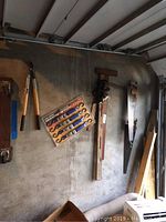 Wide view of tools hanging on a concrete wall including Fiskars lopper, ratcheting tie-downs in package, vintage bubble level, T-square, and vintage saws.