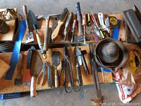 Wide view of assorted vintage hand tools laid out on wooden surface including spades, wire brushes, pruners, hatchet, clamps, scissors, paint brushes, tape measure.