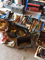 Image shows large group of tools and hardware on garage floor and workbench, including hammers, mallets, saw, sharpening stone, brush, and bags of salt.