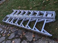 Photo showing two aluminum utility ladders lying on the grass, 8 feet in height with noticeable paint stains and general wear.