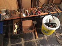 Vintage hand tools displayed on wooden shelf and plastic bucket, includes screwdrivers, wrenches, clamps, files, mallet, jumper cables