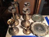 Six vintage weighted brass candlesticks and two plates on a table, showing variety in candlestick sizes and plate styles.