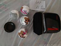 Four LOL Surprise toy balls arranged next to a small black tray and a black cooler bag closed with red zipper.