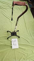 Photo of the quad cane upright on a light green surface showing the brown handle, curved shaft, and large square black base with four contact points.