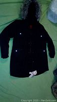 Full front view of the black winter jacket laid flat, showing hood with faux fur and multiple front pockets with buttons.