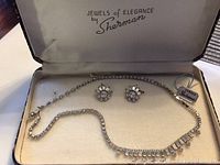Jewelry set of rhinestone necklace and matching clip-on earrings in original Sherman box seen from above showing the Jewels of Elegance by Sherman logo inside the lid.