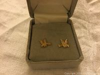 Pair of small 10 karat gold eagle shaped stud earrings inside an open small grey jewelry box on a fabric surface.