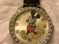 Close-up photo of watch face showing colorful Mickey Mouse character in red shorts and yellow shoes, silver dial numbers, and a bezel decorated with clear crystals.