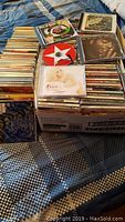Photo showing stacks of over 130 mixed music CDs on a patterned bedspread, with visible album covers including Cher, Bob Dylan, Stevie Ray Vaughan and others.