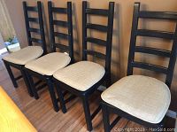 Four black IKEA Kaustby style kitchen chairs with beige cushions lined up side by side on wood floor next to light wood table.