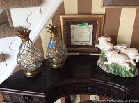Two glass pineapple accent lights, a ceramic bunny figurine, and one framed artwork on top of a dark wood table in front of striped wallpapered wall and stairs.
