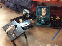 Overview of various power tools including jig saw table, Black and Decker router, Makita belt sander and circular saw in cases, DeWalt hand sander, and accessories on concrete floor.