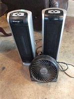 Two black and gray Lasko tower heaters with controls on top and one black Honeywell fan with spiral grill on concrete floor in garage area.