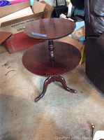 Full view of the vintage two tier wooden table standing on the floor with a dark wooden finish and three curved legs.