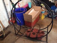 Photo showing black metal circular wood storage rack with rectangular base, some rust visible, located indoors in garage with cluttered background