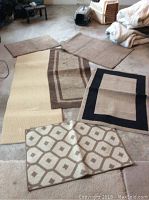 Six area rugs displayed on a carpeted floor showing variety in size, color, and pattern. Some rugs are folded or partially stacked.