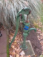 Full side view of vintage green cast iron hand water pump standing on concrete base outdoors, showing lever, spout, and mounting holes.