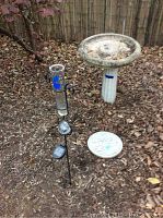 Full view of garden items showing the cement bird feeder, glass rain gauge on iron stand, and USN stepping stone placed outdoors on leaf-covered ground.