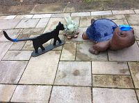 Full view of three outdoor garden decor figurines on paved patio: ceramic turtle, ceramic bunny, black iron cat boot scraper