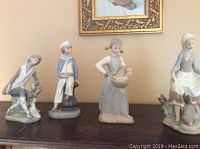 Photo of four porcelain figurines displayed on a dark wooden surface, includes two men and two women, one with kitten, others with dogs, detailed facial features and pastel colors.