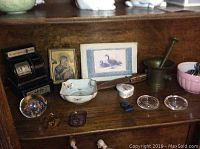 Photo showing vintage toy cash register, brass mortar and pestle, Limoges bowl with butterfly designs, blown glass paperweight, small framed religious icon, bird picture, and small glass dishes on wooden shelf.