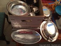 Silverplate items including oval platter, covered dishes, and bowl visible inside a cardboard box and laid out on table