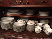Photo showing stacked Noritake Silverdale china including plates, cups, serving bowl, gravy boat, and platters placed in a wooden cabinet.