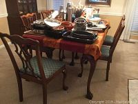 Dining table with various kitchenware items placed on top along with six chairs surrounding the table in a carpeted room