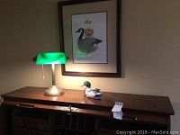 Desk lamp with green glass shade and wooden desk with seated duck decoy beneath framed dog and duck print.