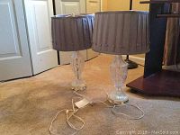 Two glass base table lamps with gray pleated fabric shades on carpet near white door.