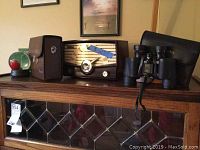 Vintage wooden Traveler radio, snow globe with golf ball, golf ball personalizer, and Bushnell binoculars with case on top of wooden cabinet