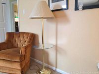 Full view of the brass floor lamp with round attached glass table beside a brown armchair.