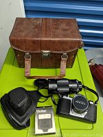 Photo of vintage Pentax film camera with attached Vivitar zoom lens, black camera case, red pouch, Vivitar flash box, and brown leather carrying case on green surface