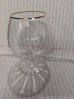 Clear brandy glass with gold rim and rounded bowl shape photographed against lined paper background.