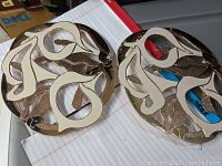 Two circular metal hot plates, bronze color with decorative cut-outs and cream swirl accents, photographed on notebook and plastic surface.
