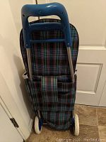 Shopping cart trolley folded and resting upright, showing metal frame with blue handle and plaid fabric bag on back side.