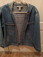 View of the front of the denim coat hanging on a hanger showing pockets and zipper.