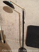 Full view of black tall standing lamp with adjustable arm, black metal shade, and power cord, standing against a wall with a shadow cast on the wall.