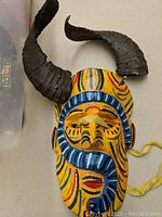 Front view of the wooden mask showing vivid hand-painted designs in yellow, blue, red, and black with large curved black horns.