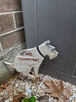 Metal dog lawn ornament shaped sitting with 'NO DUMPING!' text, showing rust and outdoor wear.