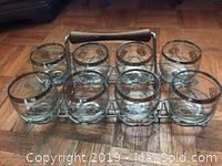 Eight clear glass tumblers set in a silver metal carrier rack displayed on wooden floor.