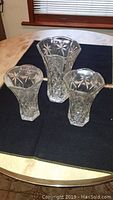 Three clear glass vases on a black cloth on a wooden table, showing intricate cut-glass star and diamond patterns with flared tops.