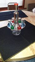 Glass decanter with silver-tone metal pump dispenser and six colored shot glasses arranged on a table.