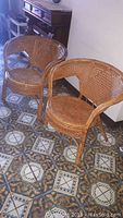 Pair of light brown wicker chairs shown from the front, displaying rounded backs and woven seats on a patterned floor.