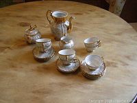 Full set of porcelain tea service including teapot, sugar bowl with lid, creamer, 7 cups on 9 saucers with gold detailing on white porcelain.