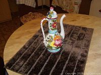 Full view of the colorful ceramic tea server on a dark striped textured rug on a round table.