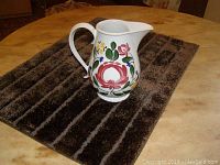 White ceramic pitcher with colorful floral decoration photographed on a dark brown textured surface.