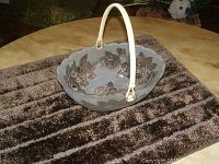 Glass fruit bowl placed on a table, showing the oval shape, frosted glass with raised flower motifs, scalloped rim, and attached white handle.