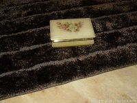 Rectangular alabaster keepsake box with floral painted lid and metal trim, photographed on textured fabric.